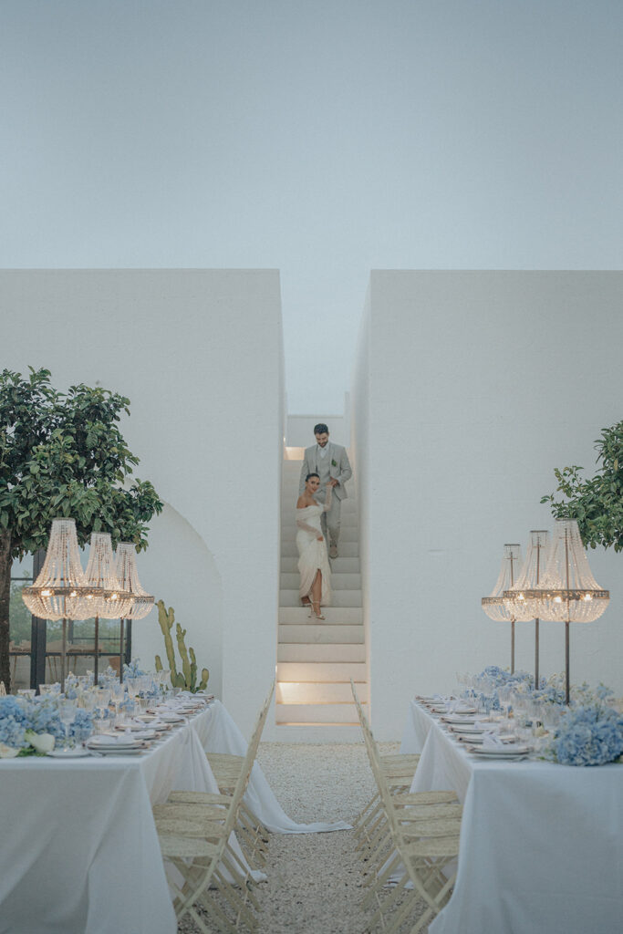 Wedding reception tables framed by white walls and stairway at Masseria Moroseta in Puglia, Italy, with chandeliers and blue hydrangea florals.