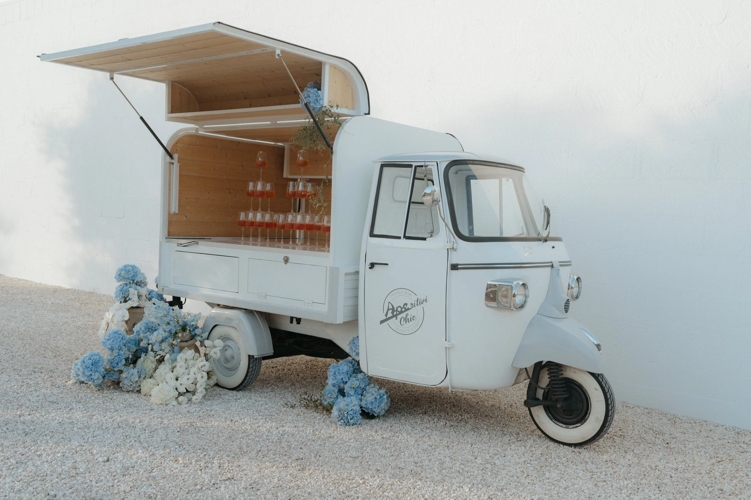 The kind of welcome your guests will never forget — an Aperitivo truck tucked into the courtyard at Masseria Moroseta, Aperol spritzes lined up, framed by soft blue blooms.