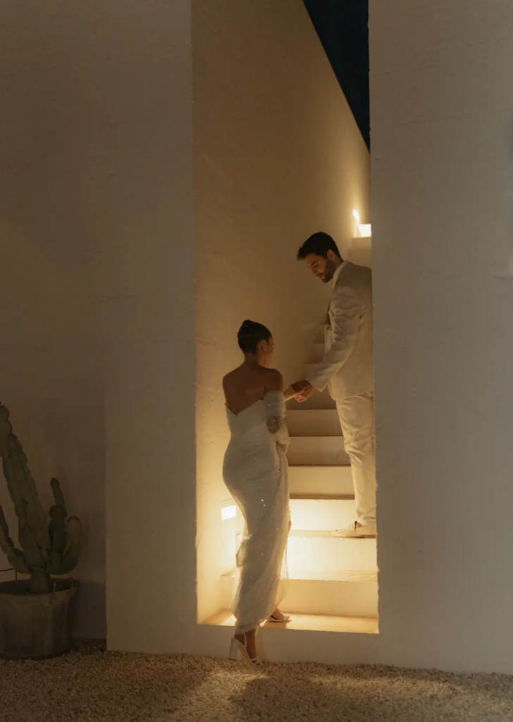 Bride in an elegant off-the-shoulder gown ascending a softly lit staircase while holding hands with the groom in a light suit at Masseria Moroseta in Puglia, Italy — captured by destination wedding photographer Suzy Anderson.