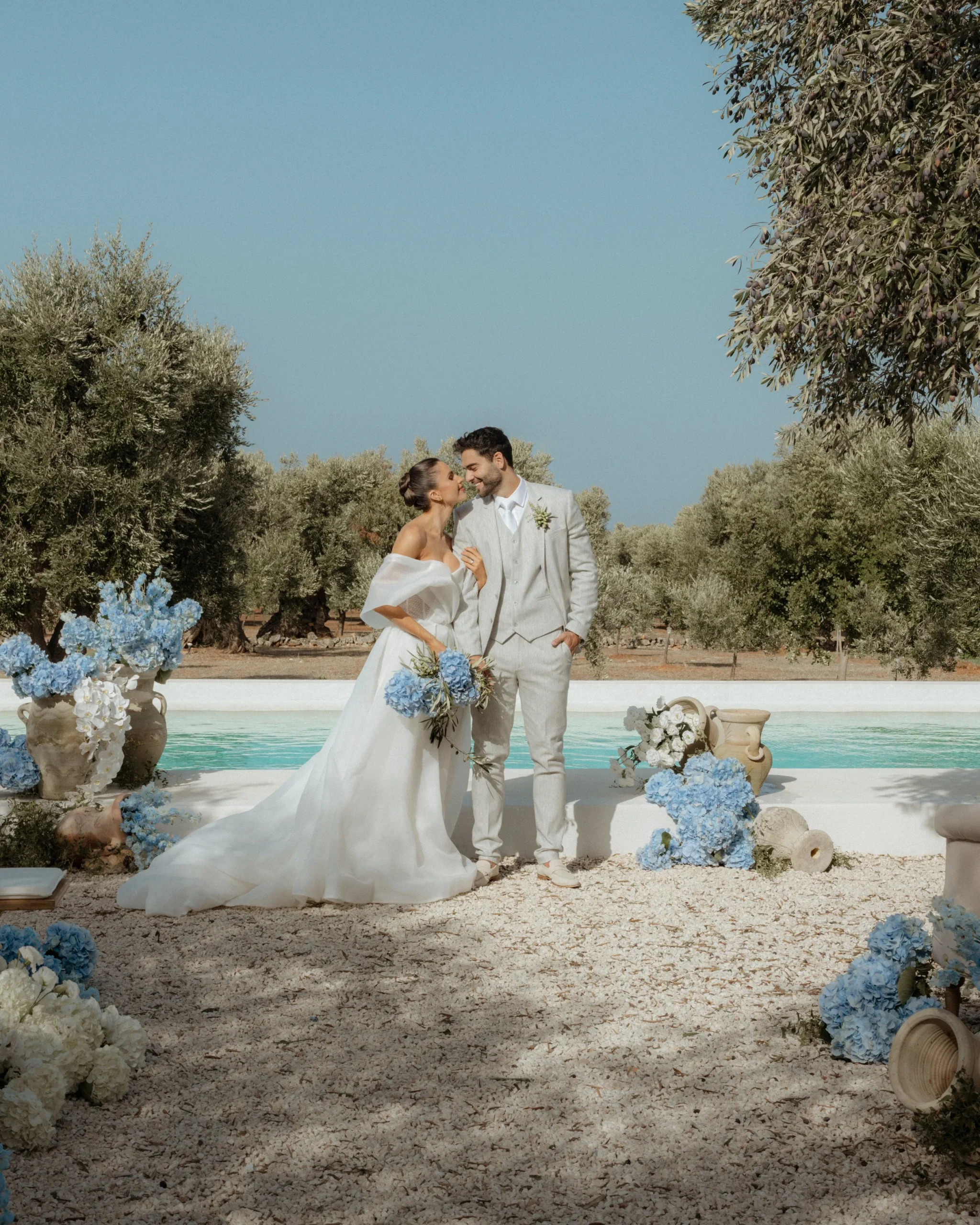 An aisle framed by olive trees, the water glistening just beyond — a ceremony that feels equal parts private and cinematic.