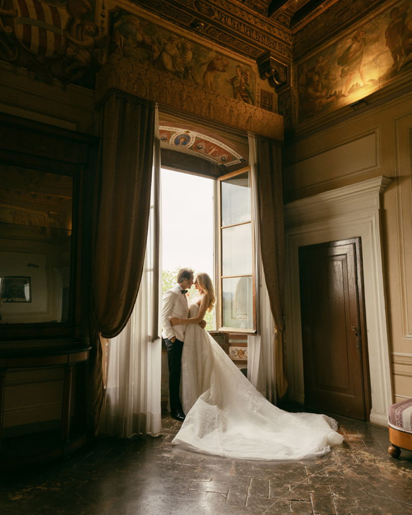Bride and groom by window at Villa Corsini Florence with soft side light, sheer curtains, and long wedding dress train in historic villa interior