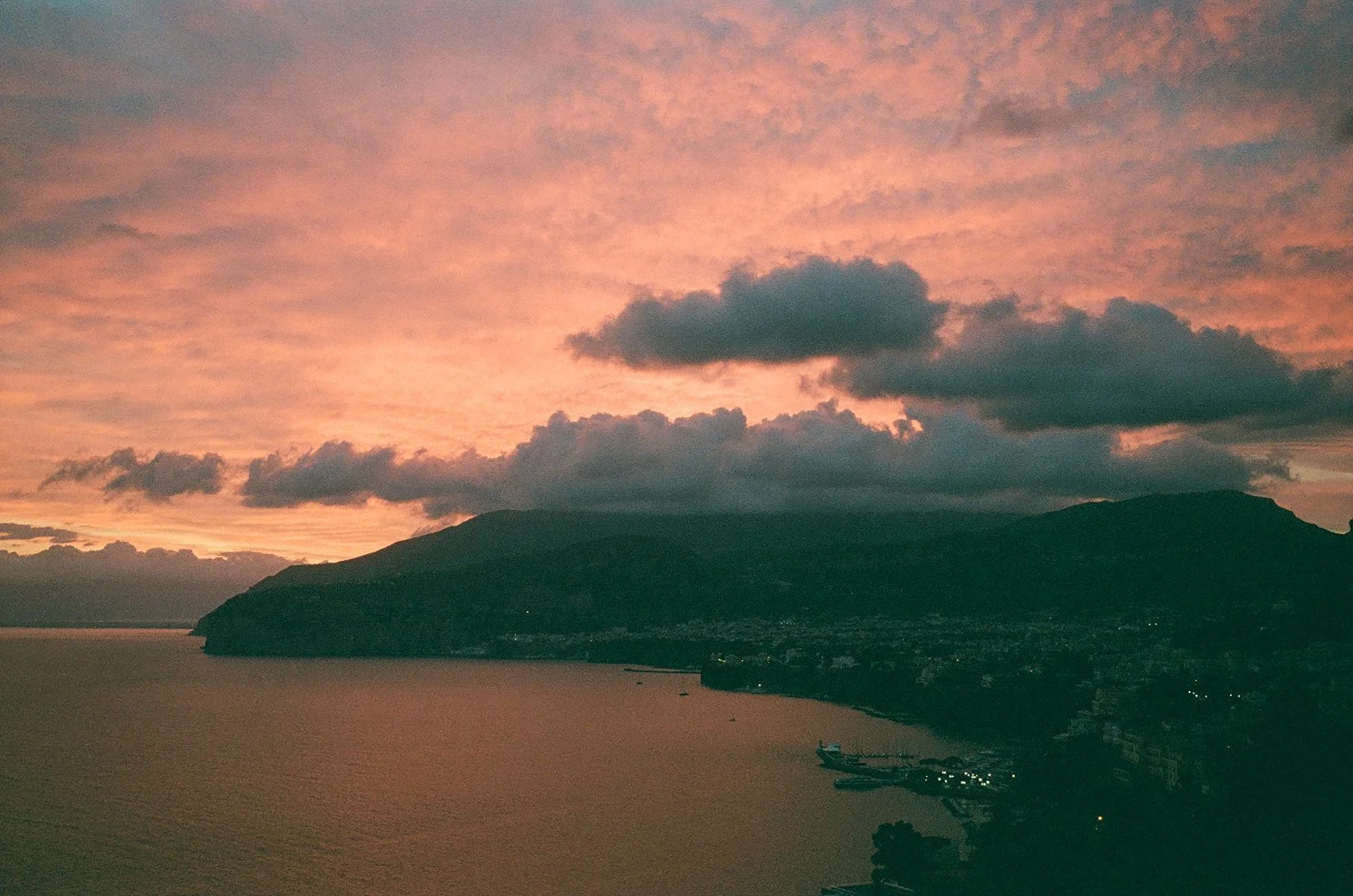  A cinematic sunset over the Amalfi Coast, where the sky blazes in soft coral and rose tones, melting into the deep indigo of the approaching night. Dark clouds drift above the silhouetted mountains, while the coastline below glimmers faintly with the first evening lights. Captured on film for a nostalgic, timeless feel. 