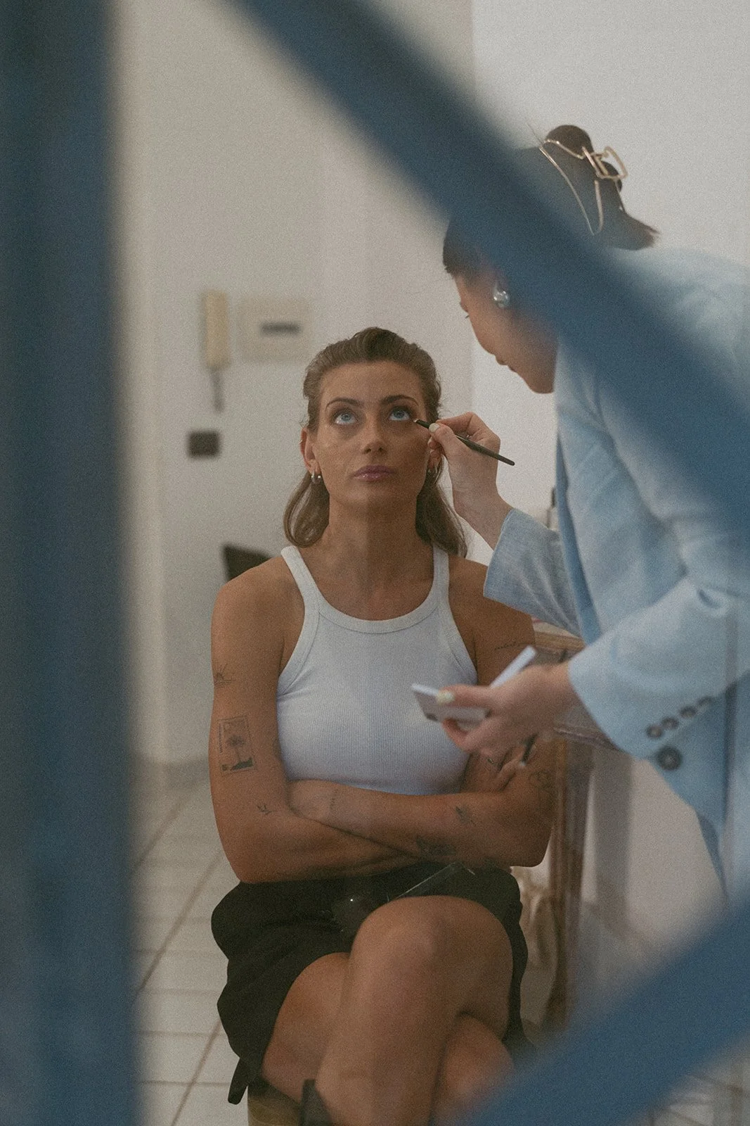  A makeup artist, Dina from  Make-up Frankfurt   carefully applie s eyeliner to a woman seated in a relaxed pose, wearing a white tank top and black skirt. The shot is framed creatively through a railing, adding an intimate, behind-the-scenes feel. 