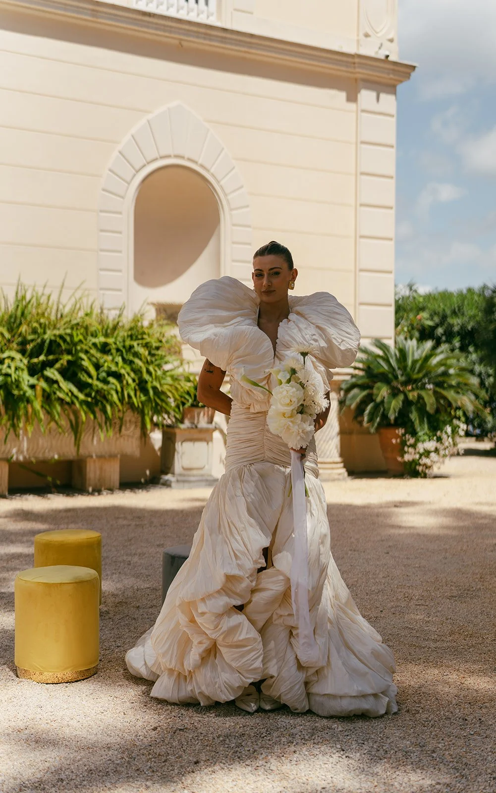  The bride stands just after the ceremony, holding her bouquet by Motif Flower. She’s wearing her Murashka gown, looking directly at the camera with a calm, natural expression. The moment feels candid and real—no posing, just her in that quiet, beautiful pause. The soft light highlights the texture of her dress and the effortless shape of the flowers, capturing something honest and true. 