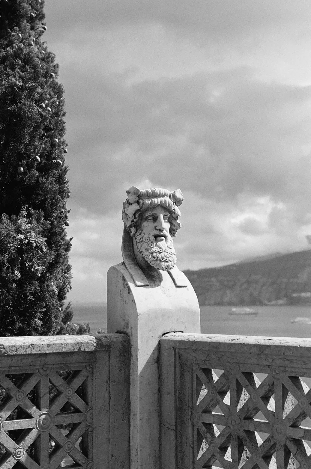  A weathered marble bust crowned with laurel stands sentinel at Villa Astor, gazing out over the Tyrrhenian Sea. The intricate stone latticework and towering cypress frame the scene, while the horizon fades into a soft, moody haze. Captured in timeless black and white for a sense of quiet grandeur.   