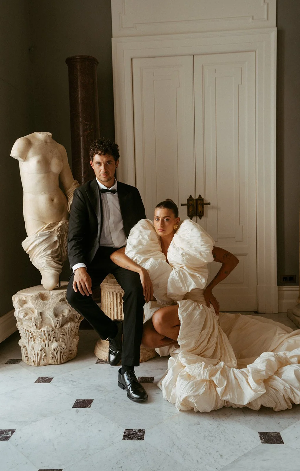  An editorial portrait of the bride and groom inside, posed next to a statue. The groom sits, the bride squats beside him. Both hold serious expressions, looking straight into the camera with quiet intensity. The mood is modern and refined—something you’d see in a Vogue spread—clean lines, strong presence, and effortless style. 