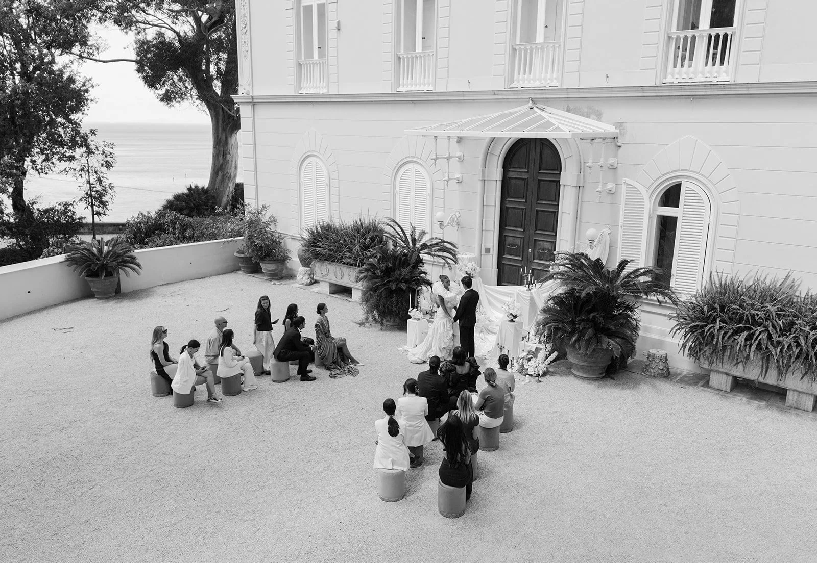  A black and white drone shot of the ceremony set just in front of Villa Astor. Guests are seated in neat rows, facing the grand doorway, where florals frame the entrance in an intentional, sculptural way. The overhead view captures the symmetry—the lines of the chairs, the shape of the villa, the balance in the design. Even without color, it feels full. Timeless, quiet, and grounded in place. 