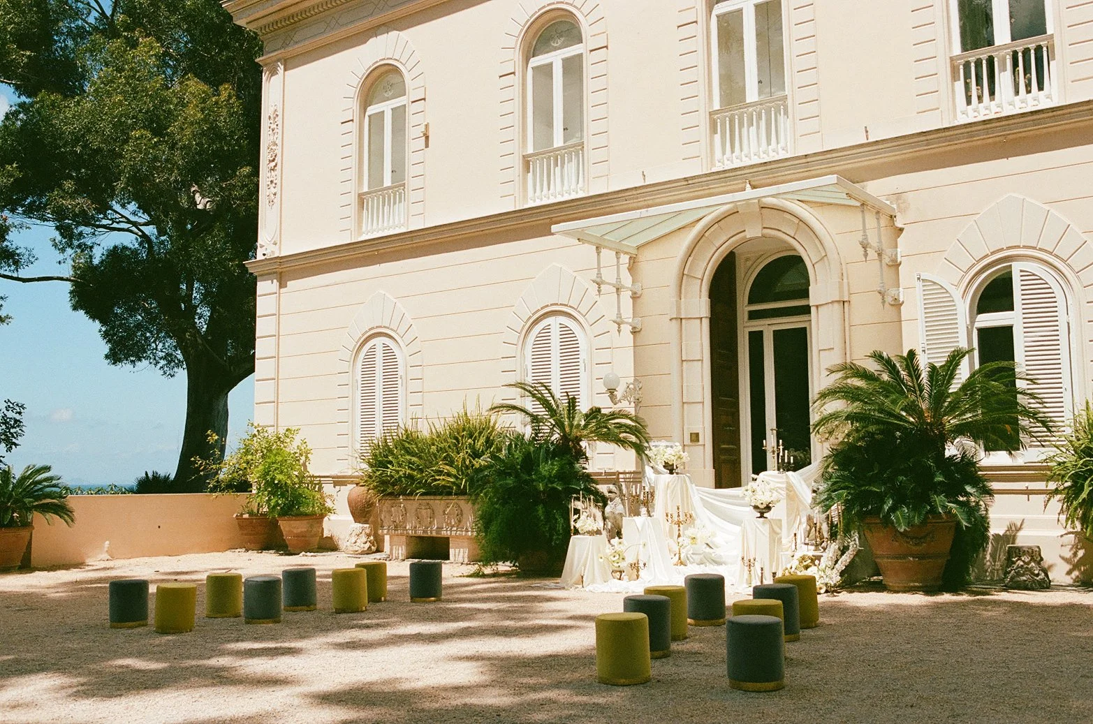  A decadent ceremony installation unfolds against Villa Astor’s grand facade — ivory silk cascading over pedestals, gilded candelabras catching the Mediterranean sun, and sculpted lions standing guard. The air feels heavy with history and romance, as if the setting itself has been waiting centuries for this moment. 