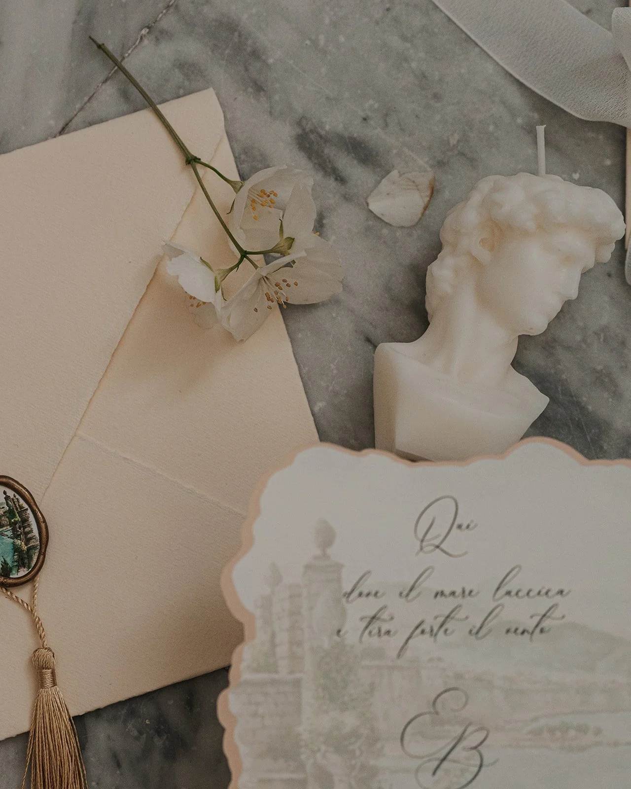  A flat lay composition in black and white featuring elegant wedding stationery with Italian script, a delicate single flower, a wax seal with a tassel, and a sculpted bust candle of David, all arranged on a marble surface. 