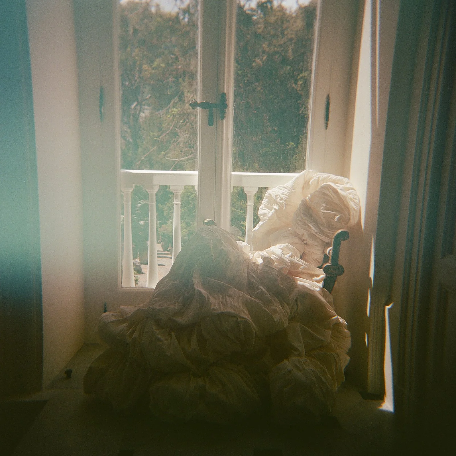  A voluminous ivory wedding gown rests on an ornate chair by the window inside Villa Astor. Soft daylight streams through the French doors, casting a romantic glow across the room. Captured on film, a subtle light leak adds dreamy color shifts along the edges, emphasizing the analog medium’s character and timeless beauty. 