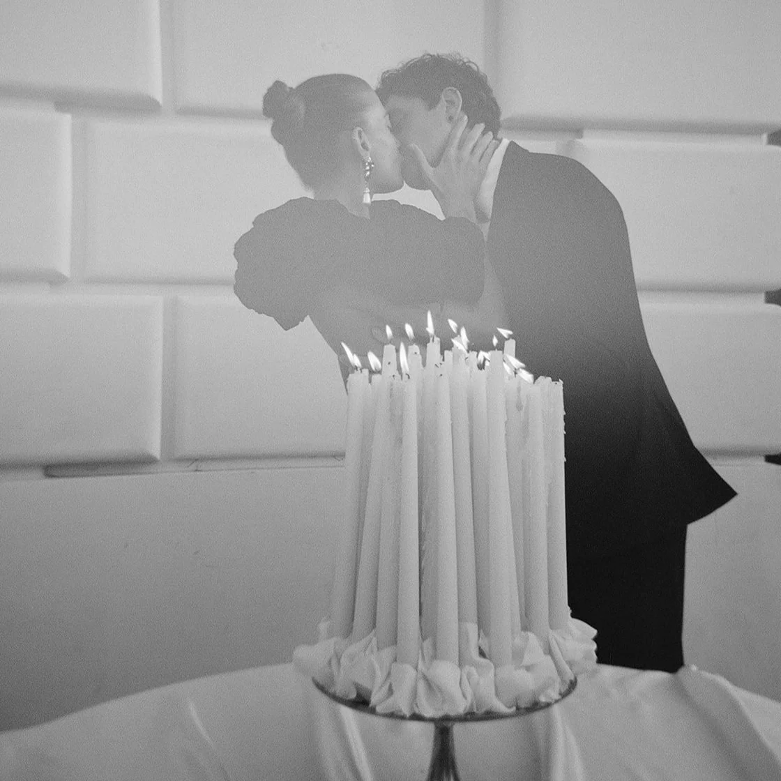  A black-and-white film photograph of a couple kissing behind a cluster of tall taper candles. The setting is architectural and refined, with sculpted white wall panels and soft, moody lighting. The bride wears elegant statement earrings and a voluminous gown, while the groom is dressed in a classic suit. The soft grain and candlelit haze evoke old-world romance and cinematic storytelling. 