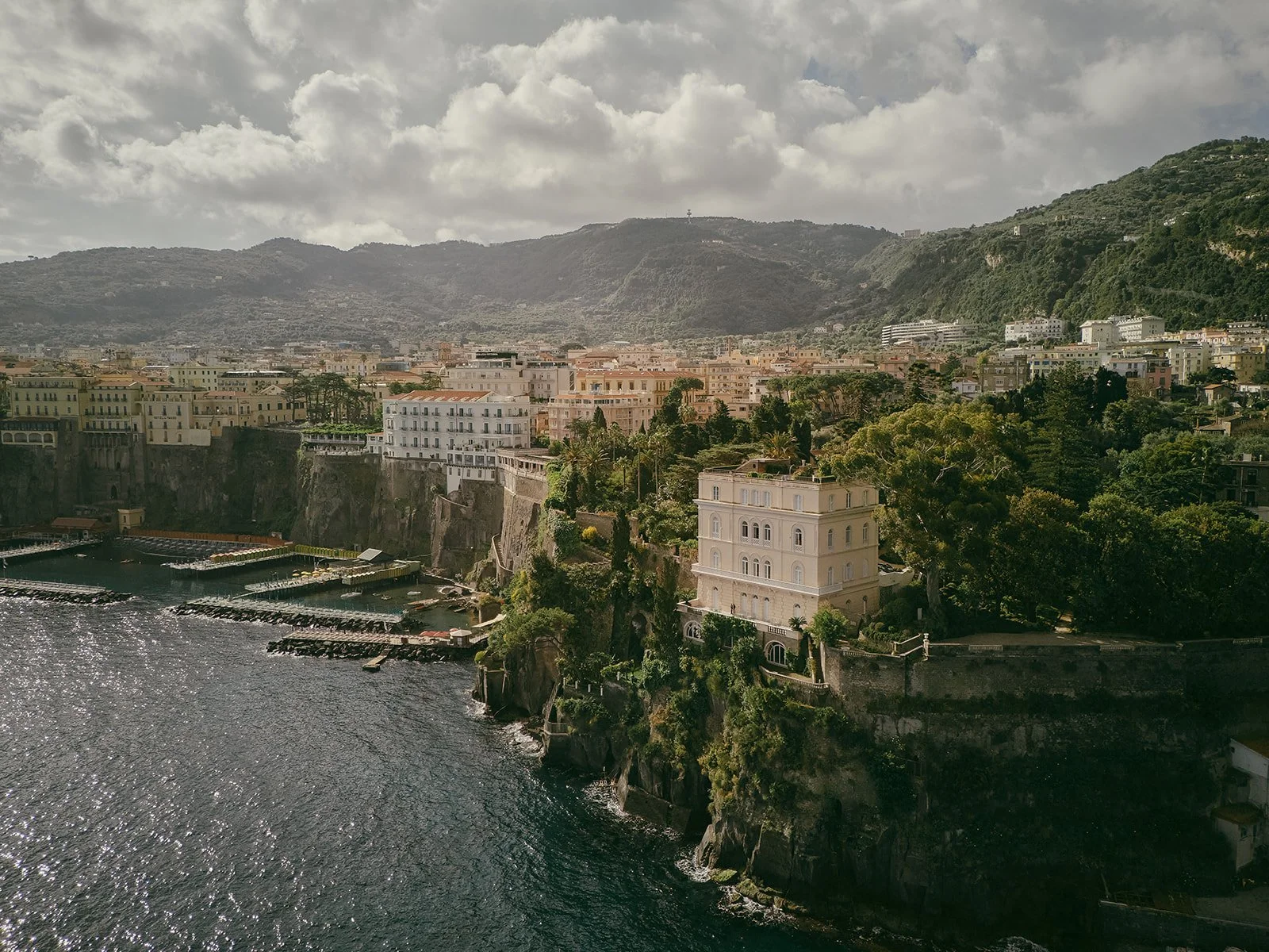  A sweeping view of Villa Astor perched atop dramatic cliffs along the Amalfi Coast, bathed in the warm, golden light of late afternoon. The symmetrical neoclassical architecture stands out against lush greenery and rugged stone, with the Mediterranean Sea gently crashing below. This cinematic moment captures the grandeur and setting that make destination weddings in Italy unforgettable. 