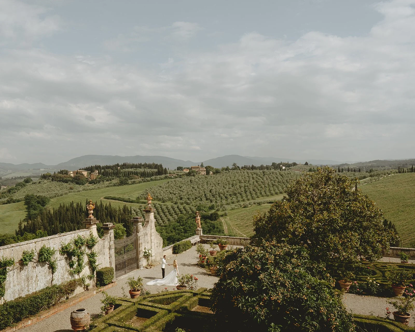 A wide-angle view of Villa Corsini's manicured garden terraces in Tuscany, Italy, with a couple standing near the ornate stone gate. The bride wears a flowing gown with a long train, while the groom stands beside her in light, neutral attire. Rolling hills filled with olive groves and cypress trees stretch out into the distance under a lightly clouded sky. The composition balances architectural elegance with expansive, cinematic landscape. 