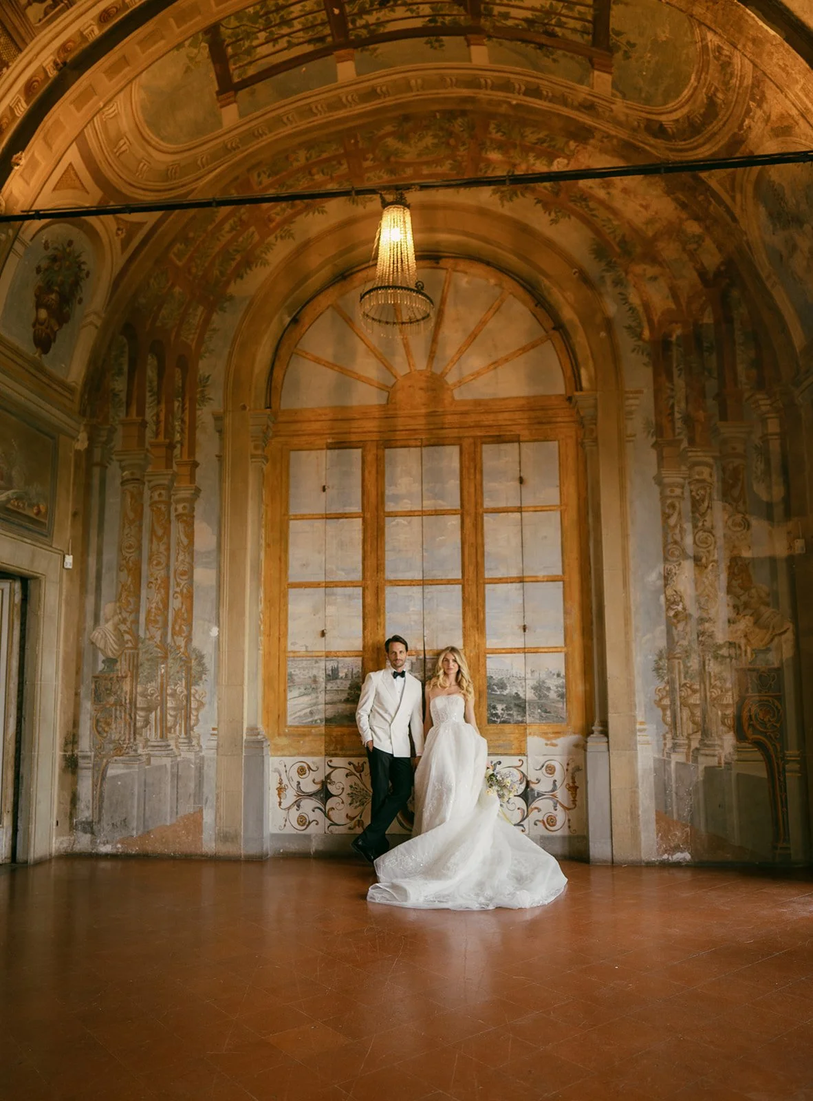  A bride and groom stand side by side inside an opulent frescoed hall at Villa Corsini in Tuscany, Italy. The groom wears a white dinner jacket and black bow tie, while the bride is dressed in a strapless ballgown with delicate embroidery. They pose beneath an arched ceiling painted with classical architectural details and a large trompe-l'œil window mural. The space feels both regal and artistic, with warm tones and dramatic lighting that add to its editorial quality. 