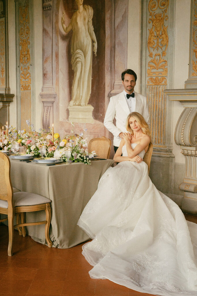 A Tuscany Wedding Styled at Villa Corsini: Timeless Editorial Inspiration in Italy Editorial wedding portrait inside Villa Corsini in Florence, Italy featuring a bride seated beside a candlelit reception table and groom standing beneath classical frescoes and sculpture.