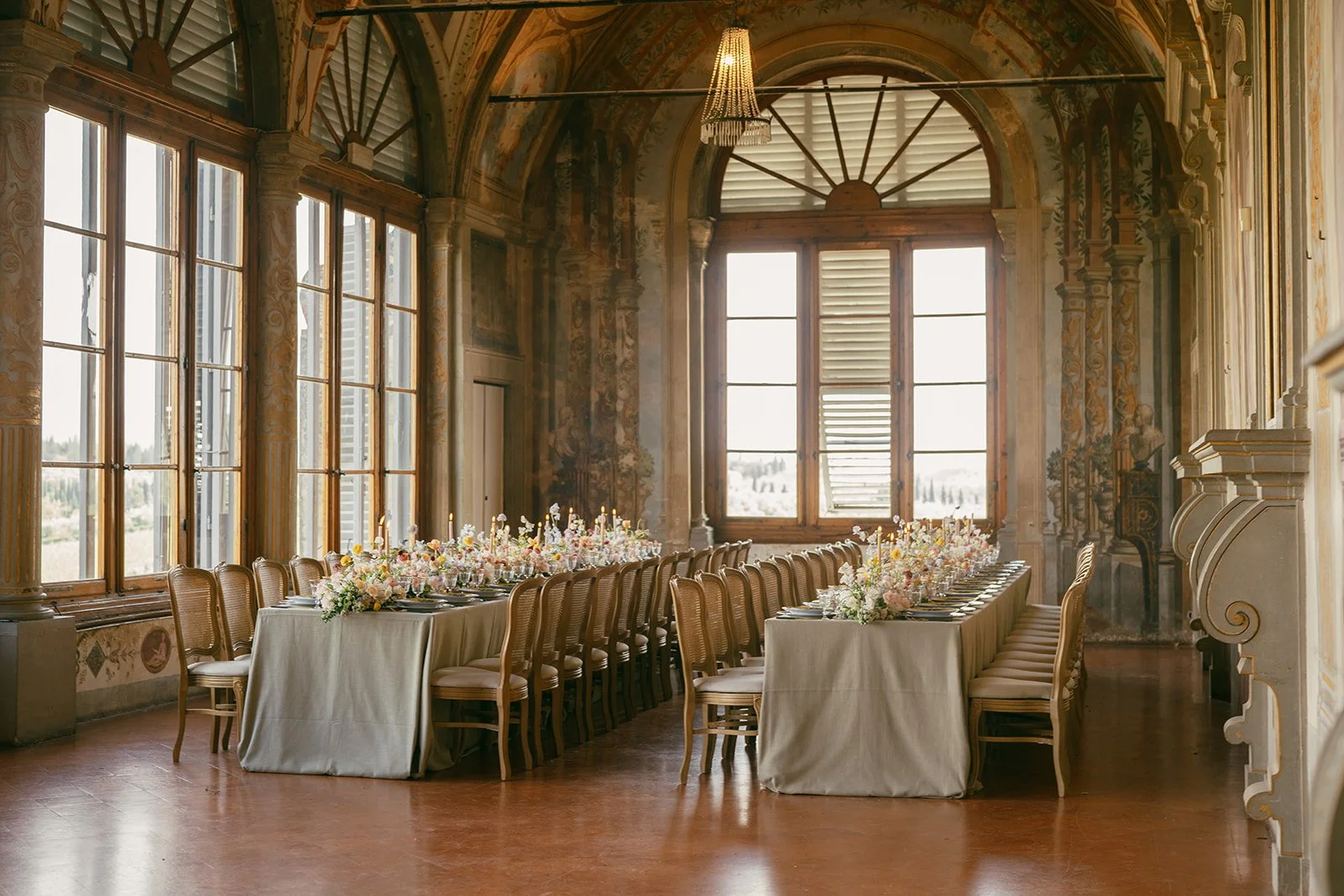  An elegant long-table wedding reception setup inside Villa Corsini’s frescoed hall in Tuscany. Neutral-toned linens cover rectangular tables lined with cane-backed chairs from Preludio Divisione Noleggio. The tables are styled with pastel floral arrangements, tapered candles, and layered place settings beneath soaring windows that fill the room with soft natural light 