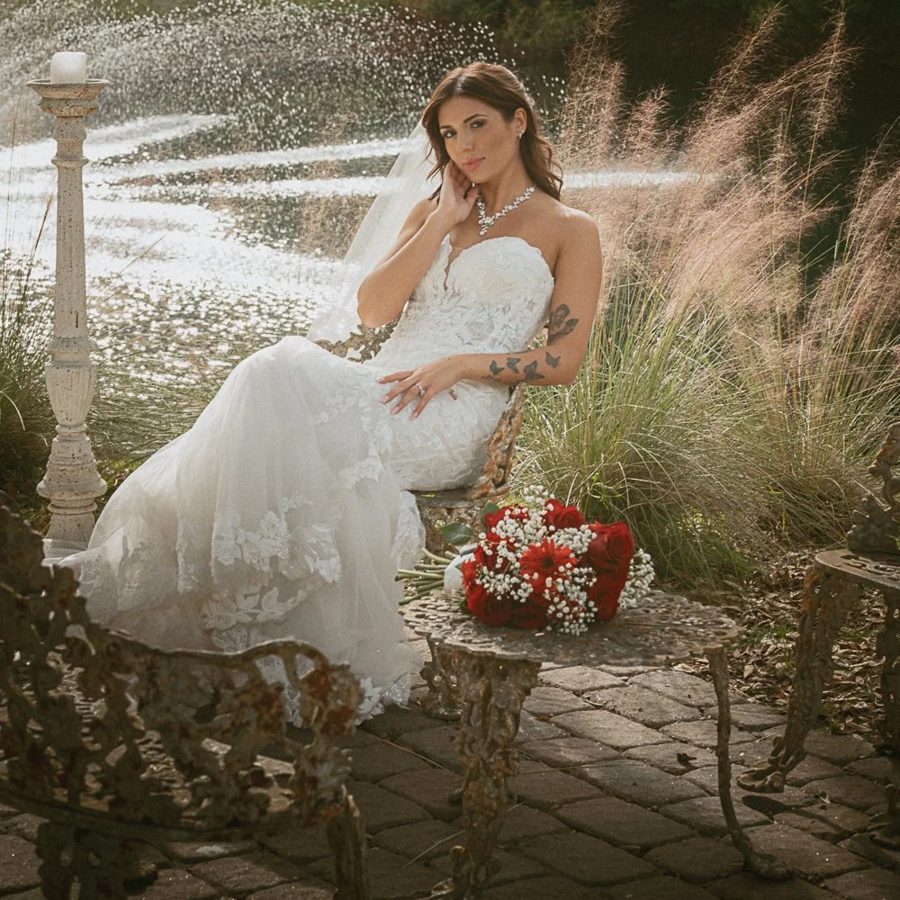 Bride in white lace wedding dress sitting on ornate metal chair by a pond, with red rose bouquet on a table, surrounded by tall grass and a candle on a stand.