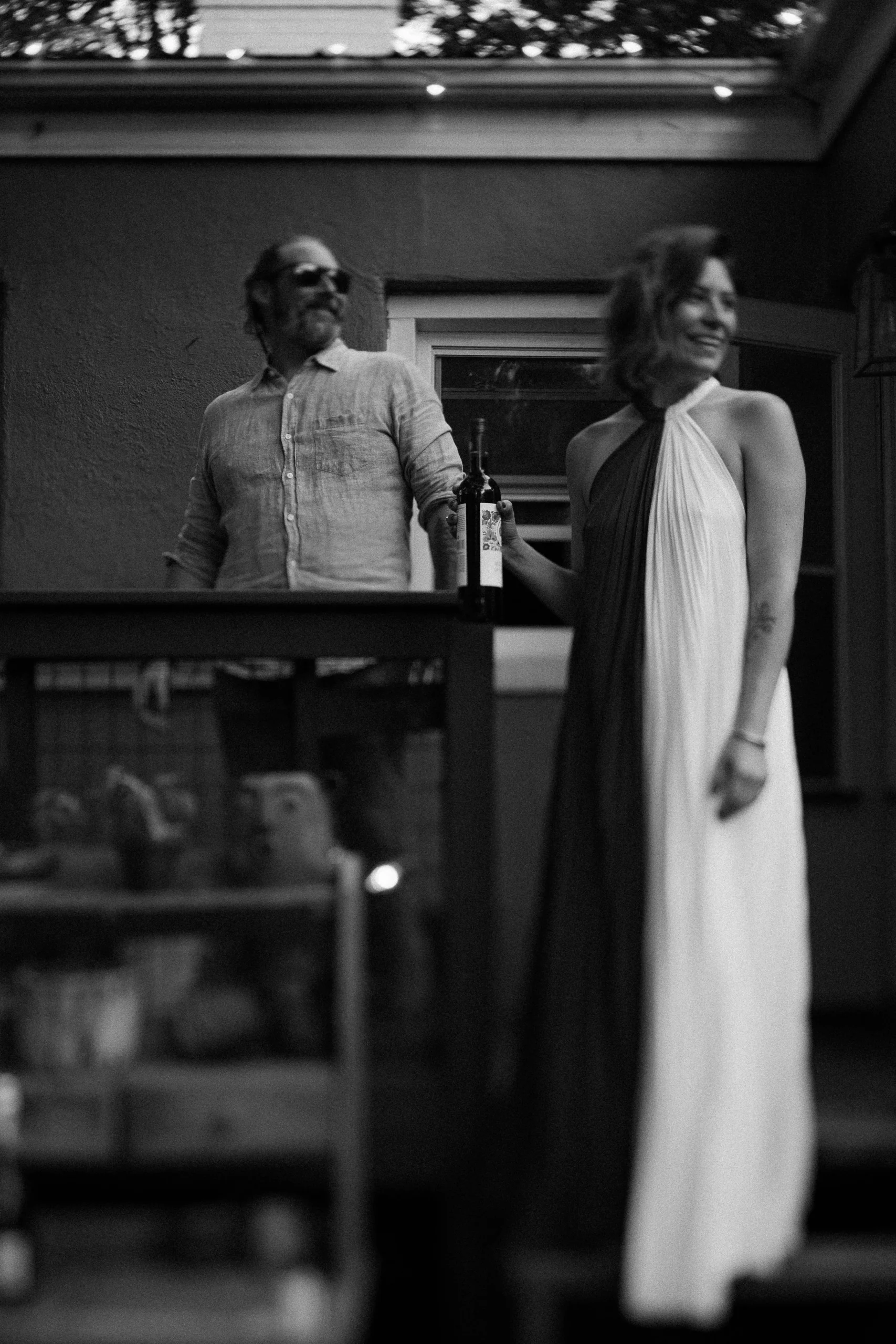 A black and white photo of a man wearing sunglasses and a woman in a two-toned dress on a porch. The woman is holding a wine bottle.