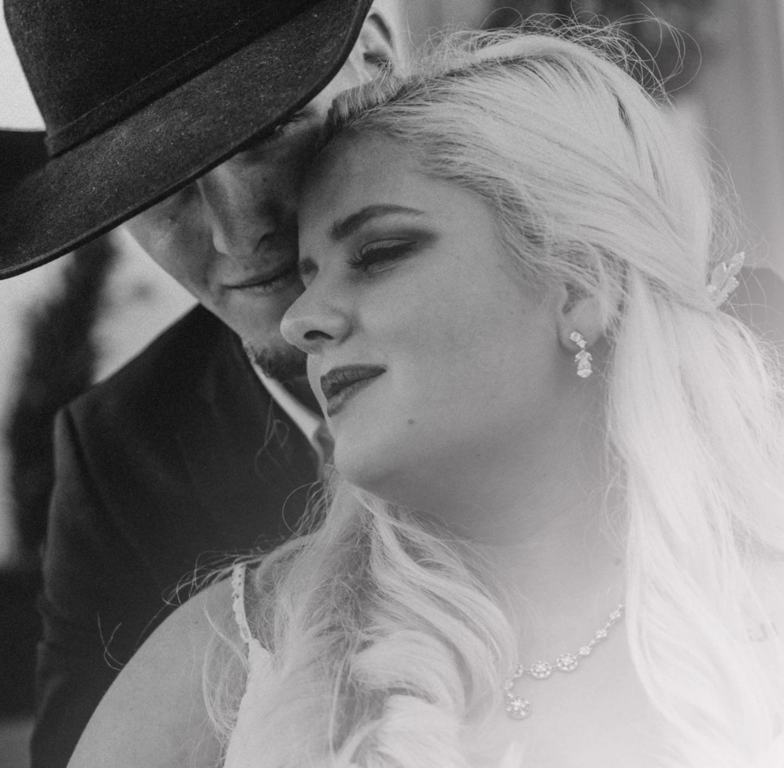 Black and white photo of a couple close together, man wearing a cowboy hat and woman with long blonde hair, necklace, and earrings.