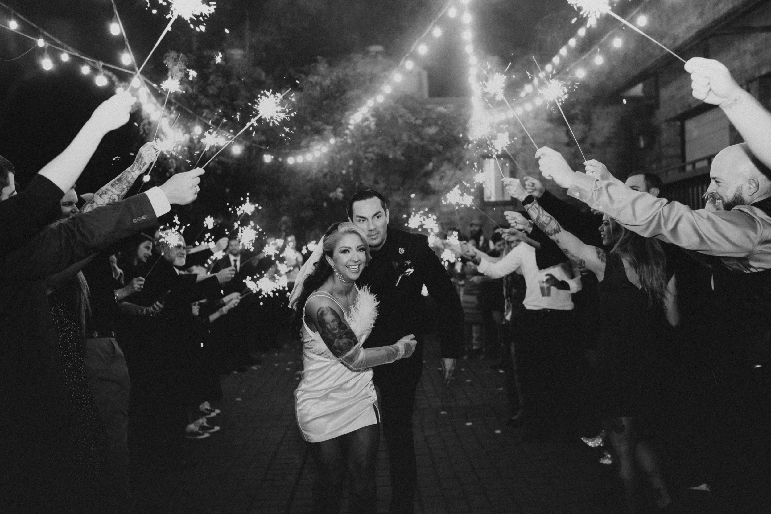 Black and white photo of a smiling couple walking through a crowd holding sparklers, under string lights, during a nighttime celebration.