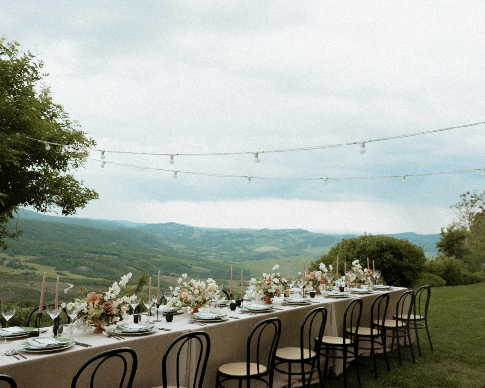 Outdoor table setting with floral arrangements and string lights overlooking rolling hills.