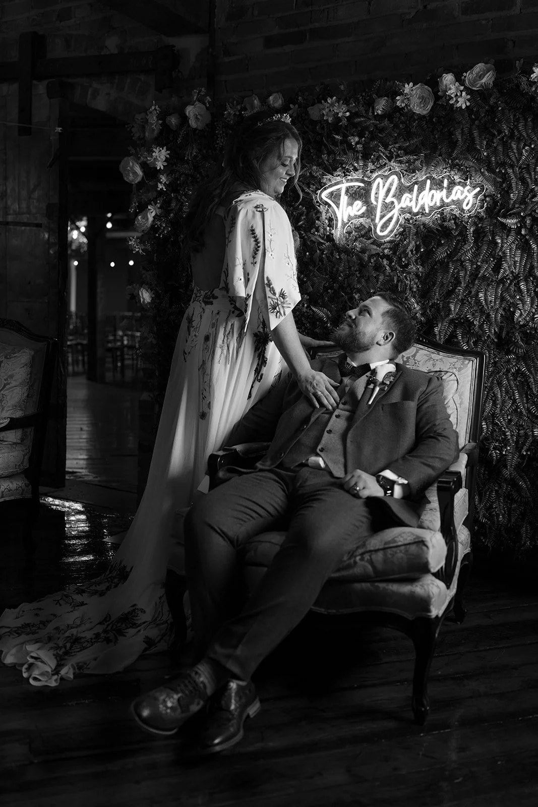 A woman in a floral dress stands next to a seated man in a suit, both in front of a neon sign reading 'The Baldrias' on a floral backdrop. The setting appears to be an indoor venue with wooden flooring.