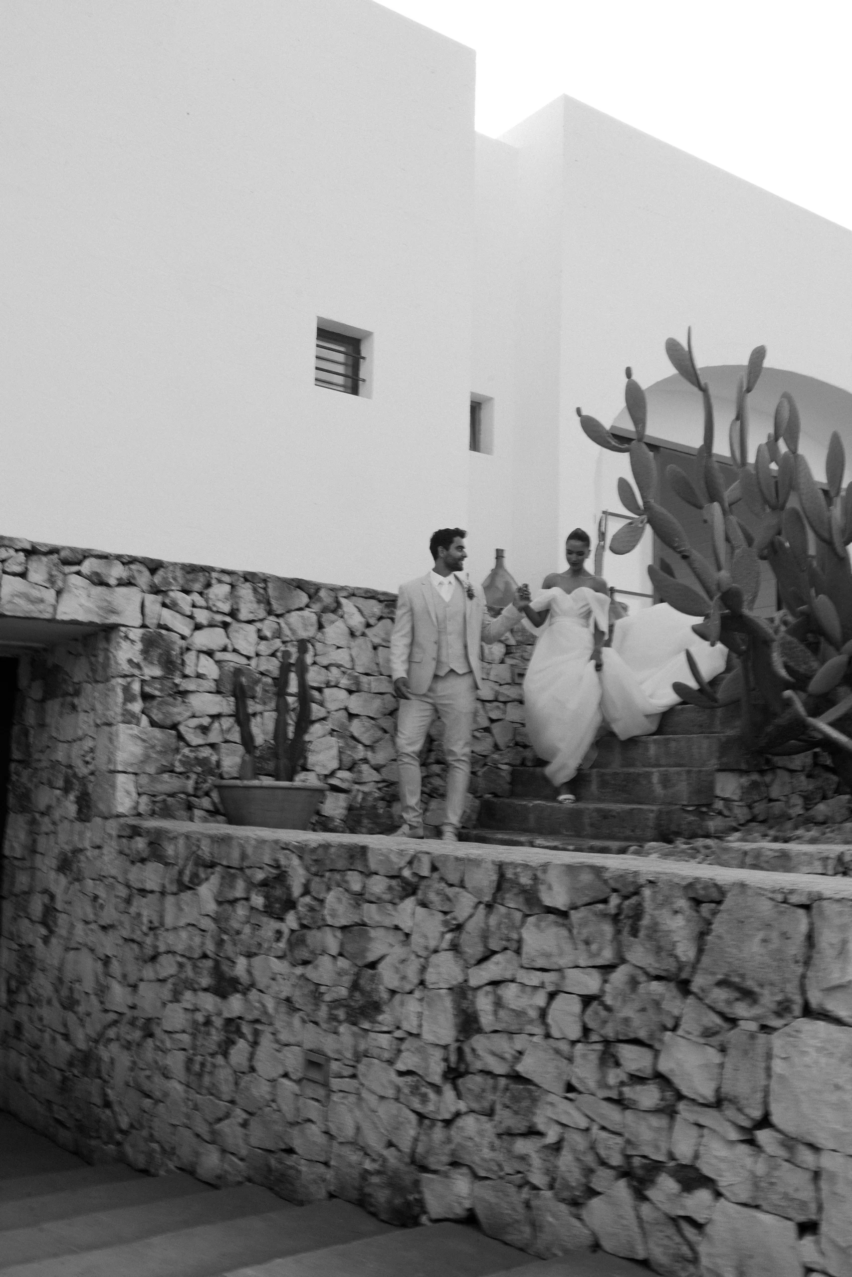 Black and white photo of a couple in wedding attire walking down stone steps beside a modern building and large cactus plants.