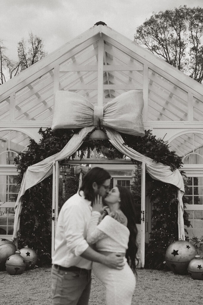 Black and white photo of a couple kissing in front of the holiday-decorated exterior of Fleurish Greenhouse in Charlotte, NC, with a large bow and greenery above the entrance.