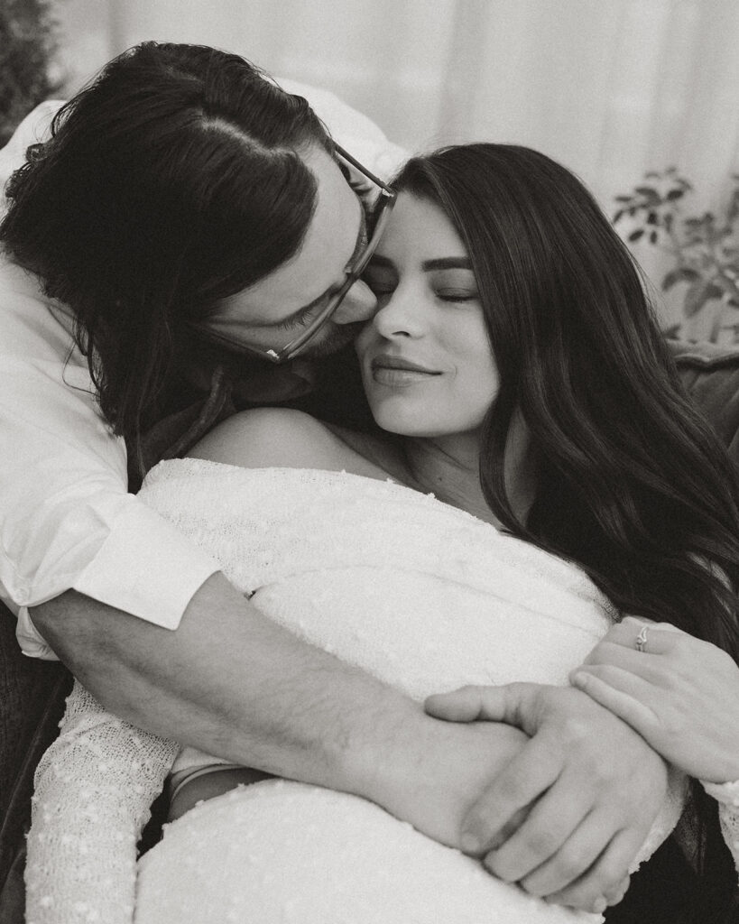 Close-up black and white photo of a couple sitting together, with the man leaning in toward the woman as she closes her eyes, taken at Fleurish Greenhouse in Charlotte, NC.