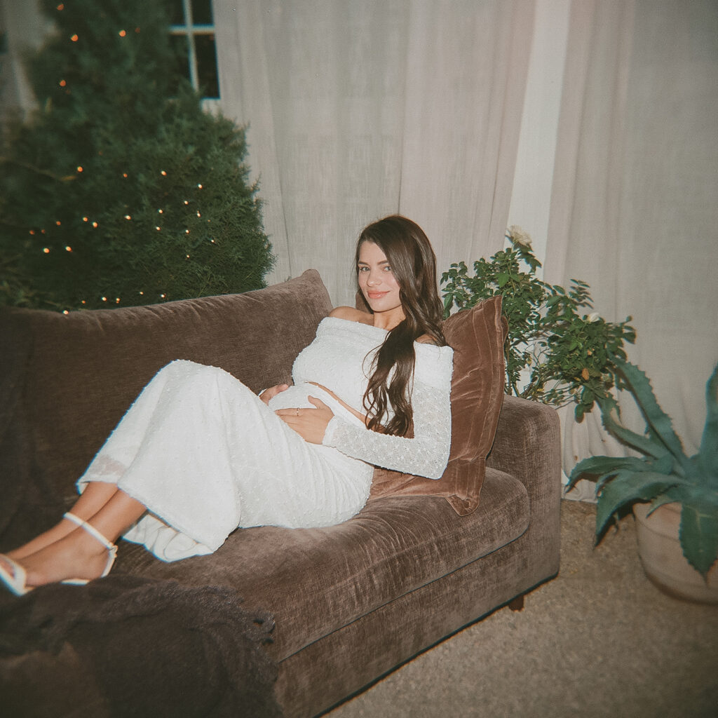Pregnant woman reclining on a brown velvet sofa inside Fleurish Greenhouse in Charlotte, NC, wearing a white gown with holiday greenery in the background.