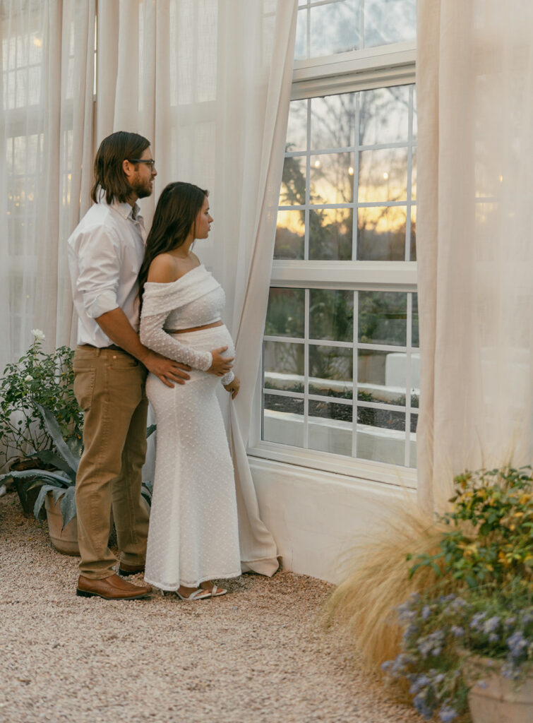 Couple standing inside Fleurish Greenhouse in Charlotte, NC, looking out a large window at sunrise; the woman wears a white dress.