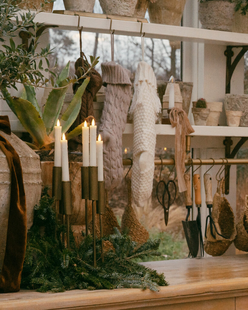 Holiday décor inside Fleurish Greenhouse in Charlotte, NC, featuring lit taper candles, evergreen branches, knit stockings, garden tools, and potted plants.