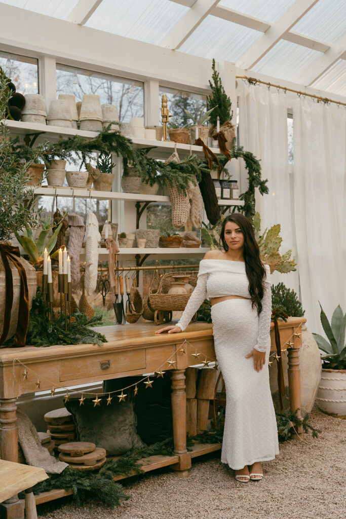 “Pregnant woman standing inside Fleurish Greenhouse in Charlotte, NC, posing beside a wooden table decorated with holiday greenery and neutral décor.