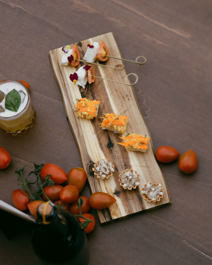 Italian appetizers on a wooden serving board surrounded by vine tomatoes and a cocktail, captured during a Monteverdi Tuscany wedding welcome dinner.