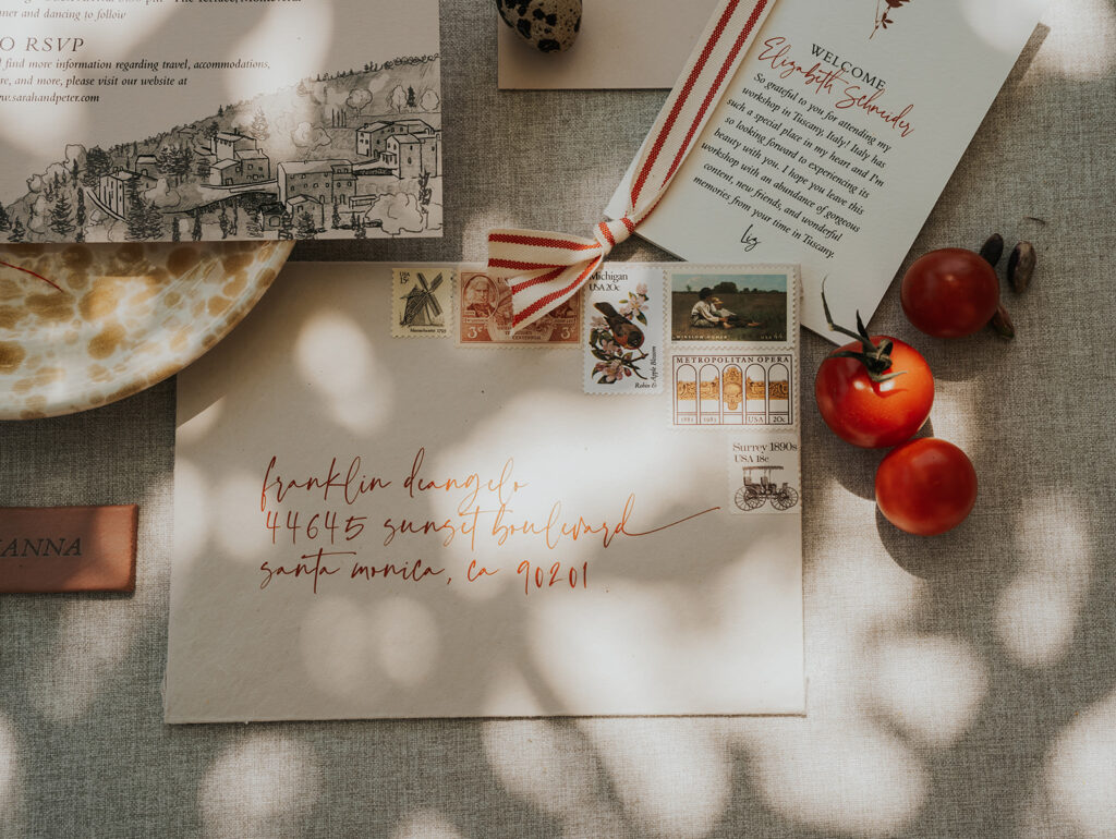 Flat lay of Monteverdi Tuscany wedding invitation suite with vintage stamps, handwritten calligraphy envelope, and fresh tomatoes in soft afternoon light.