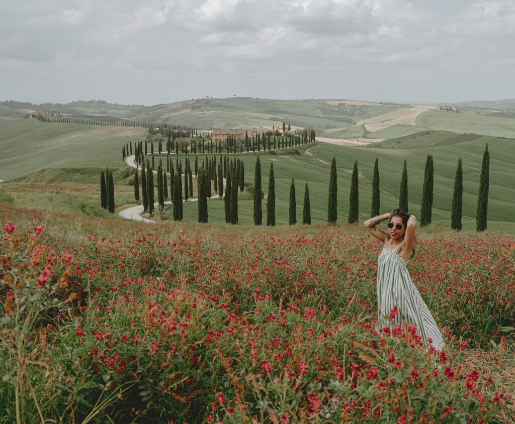 Suzy Anderson standing in a wildflower field in Val d’Orcia, Tuscany, surrounded by cypress trees and rolling green hills.