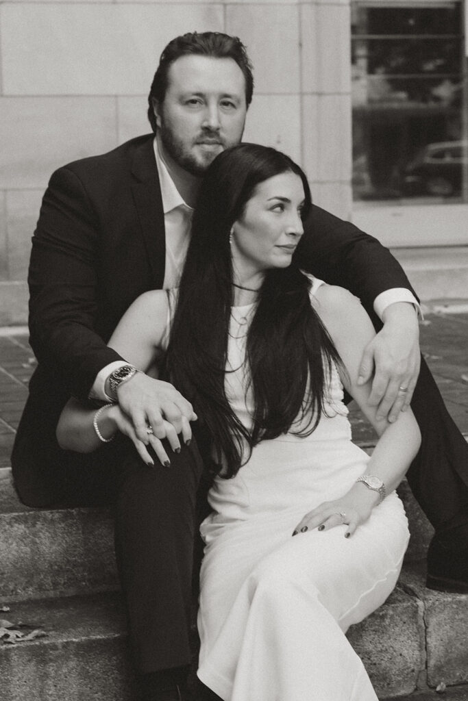 Couple sitting closely on stone steps, the man’s arm wrapped around the woman as they look in opposite directions, captured in a timeless black-and-white editorial portrait in Charlotte.