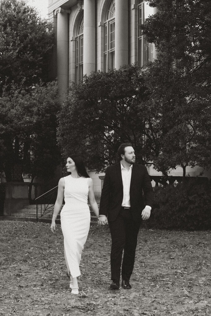 Couple walking hand in hand in front of a grand historic building, dressed in classic black and white attire during an editorial film photography session near Charlotte.