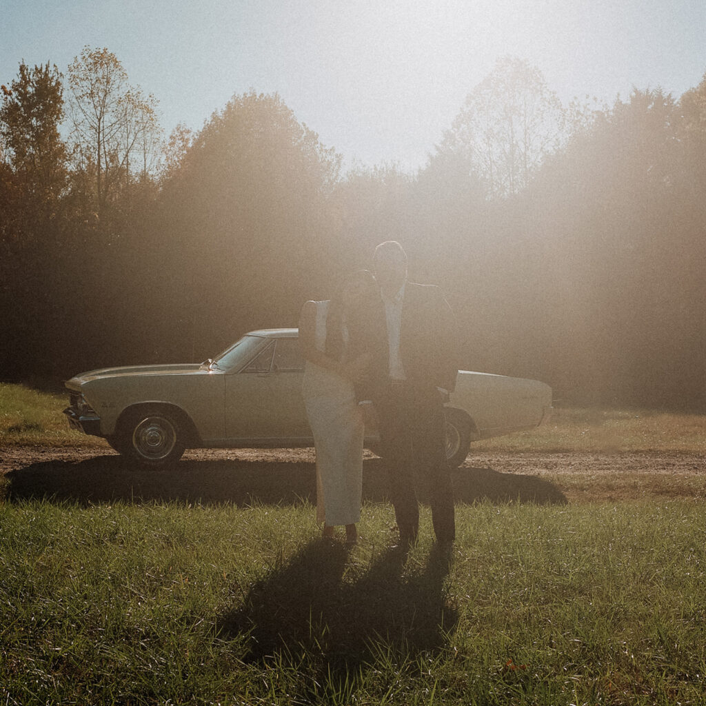 Couple standing in front of a vintage cream Chevelle at golden hour, surrounded by soft sunlight and haze during an editorial film session near Charlotte.