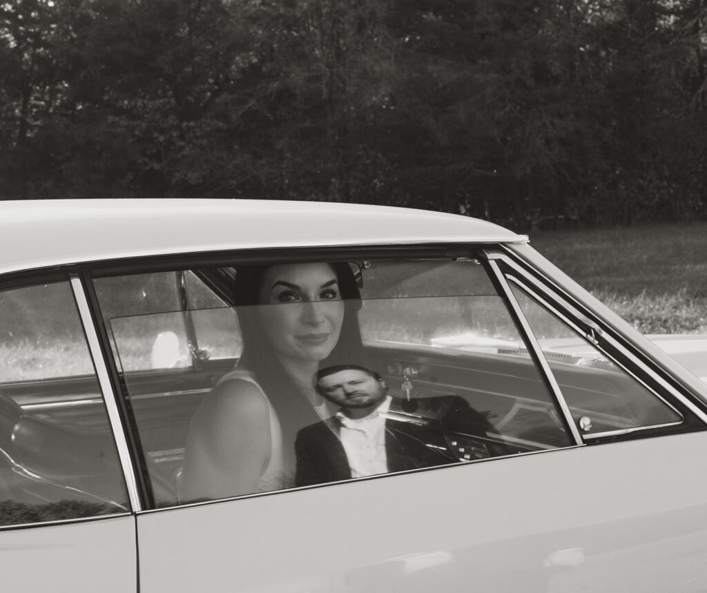 : Reflection of a man in a car window as a woman looks through the glass, captured in black and white on film.