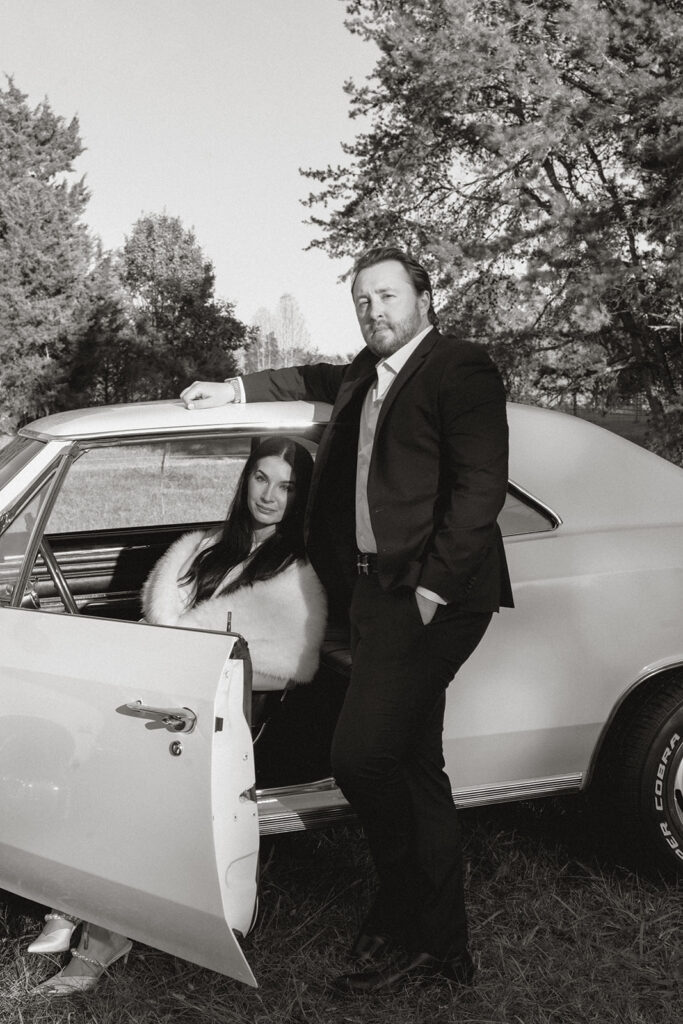 Couple posing with a vintage Chevelle, the woman seated gracefully inside the car and the man standing beside her in a tailored suit, captured in timeless black and white near Charlotte.