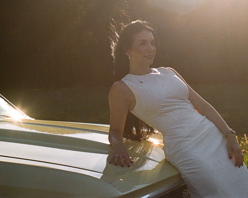 Woman in a white dress leaning against a pale yellow vintage Chevelle at golden hour, captured in cinematic film tones during an editorial couples session near Charlotte.