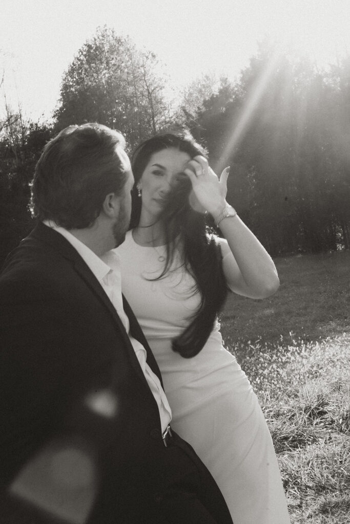 Couple sharing a quiet, sunlit moment in a field, the woman brushing her hair from her face, captured in a candid editorial black-and-white photo near Charlotte.