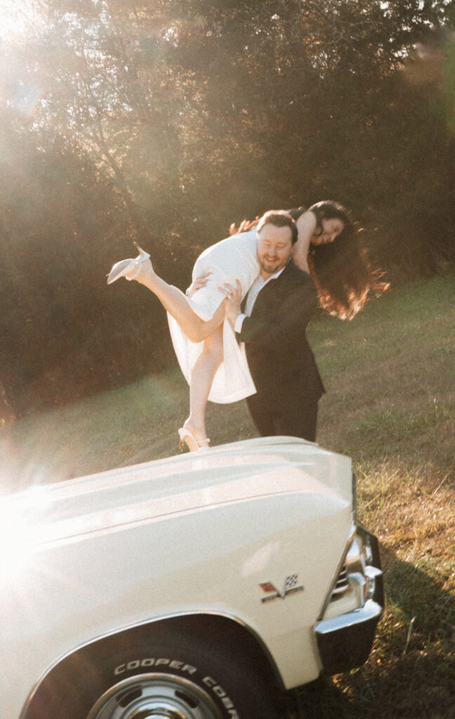 Man lifting his partner playfully beside a pale yellow vintage Chevelle in a sunlit field, captured in cinematic light during an editorial couples session near Charlotte.