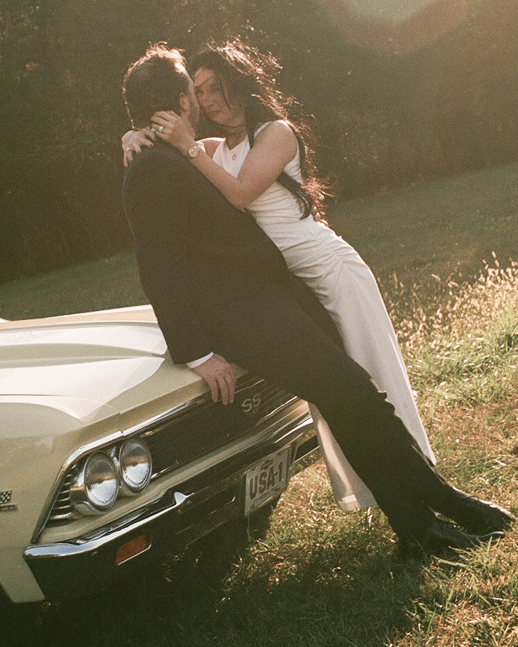 Couple embracing on the hood of a pale yellow vintage Chevelle in a sunlit field, captured in cinematic film tones during an editorial couples session near Charlotte.