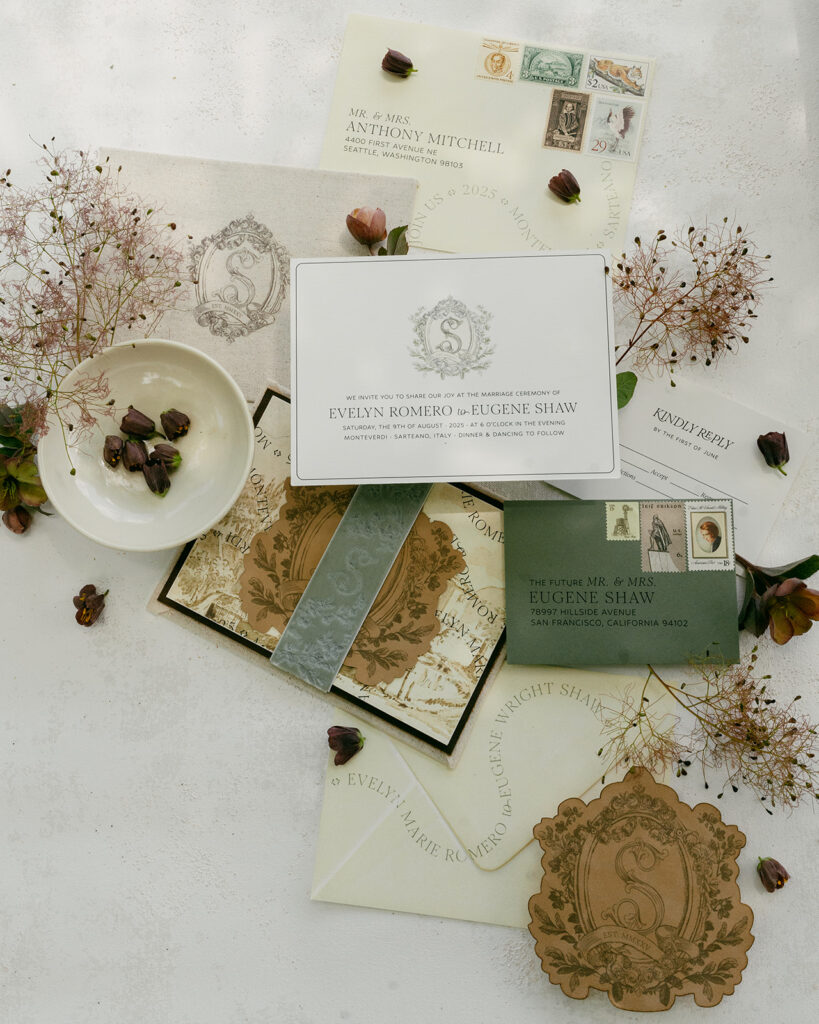 Editorial flat lay of Monteverdi Tuscany wedding invitation suite with crest monogram, vintage stamps, and hand-torn paper details.