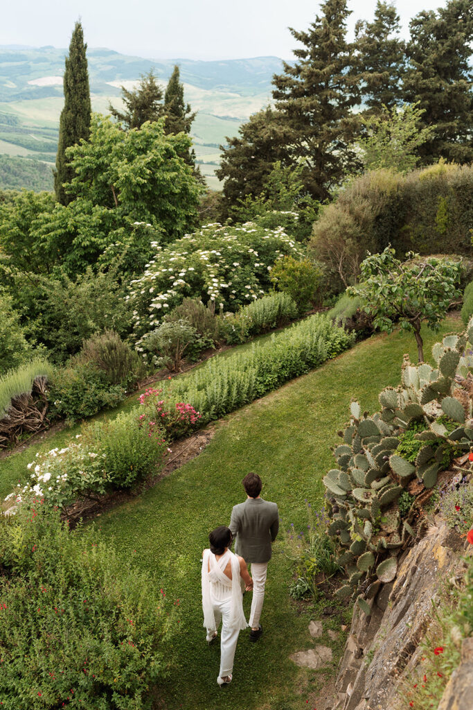 Couple walking through the lush gardens of Monteverdi Tuscany surrounded by olive trees, cactus, and wildflowers.