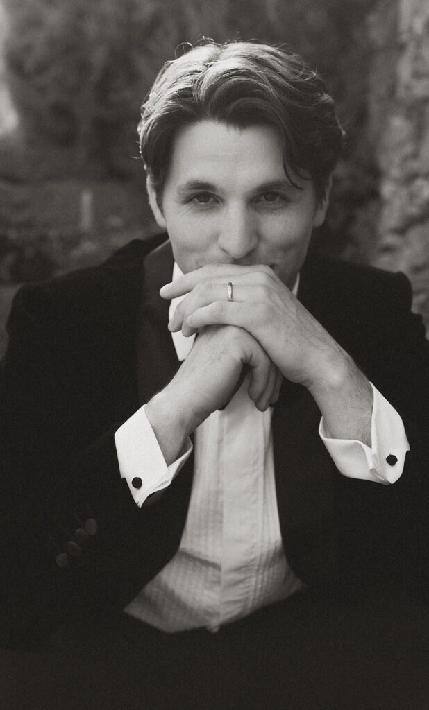 Black and white close-up portrait of the groom in a velvet tuxedo, hands folded thoughtfully, captured at Monteverdi Tuscany.