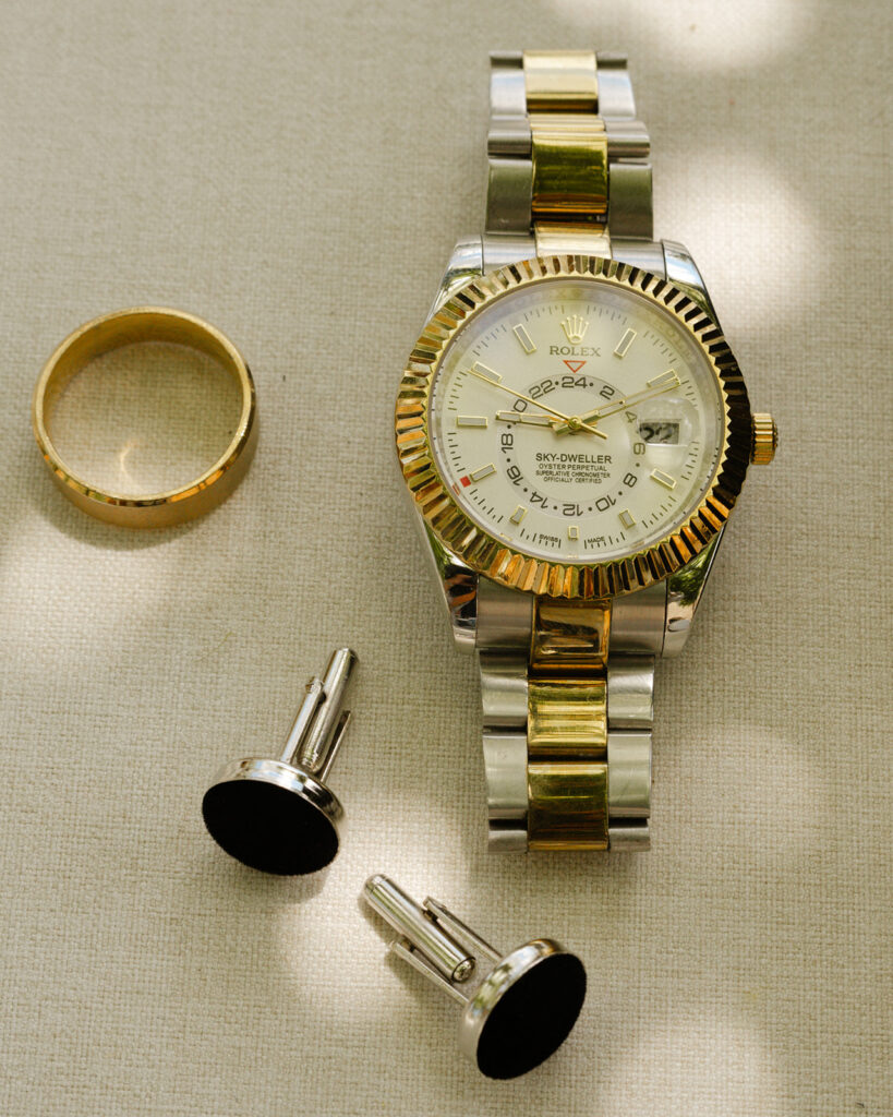 Flat lay of groom’s wedding details at Monteverdi Tuscany featuring Rolex watch, gold wedding band, and cufflinks under soft dappled light.