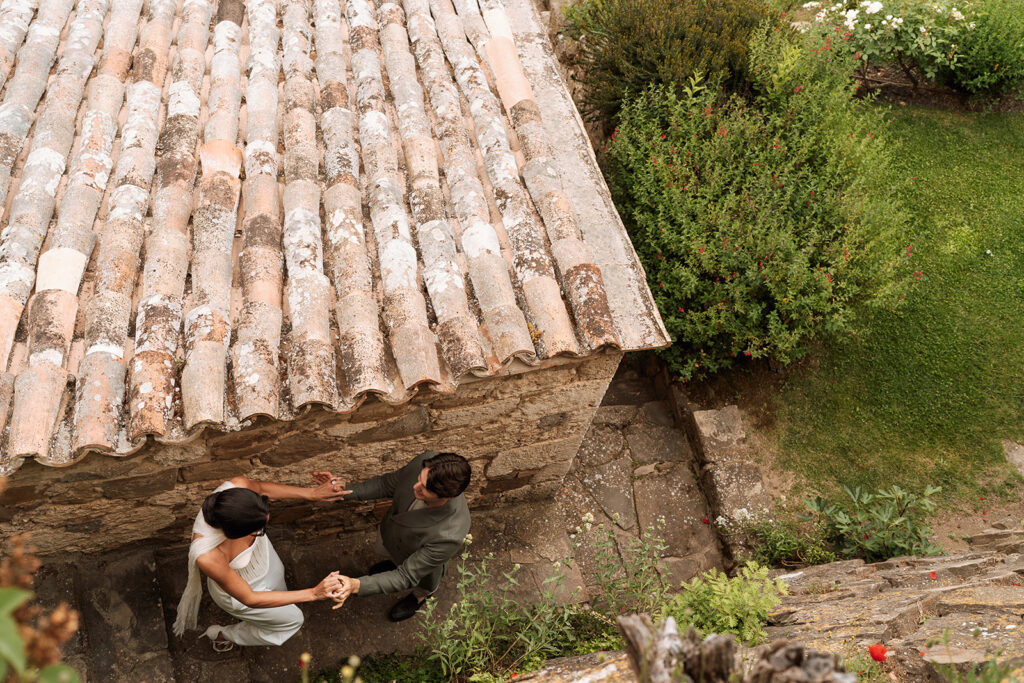 Overhead view of a couple holding hands between stone walls at Monteverdi Tuscany, surrounded by terracotta rooftops and lush garden greenery.